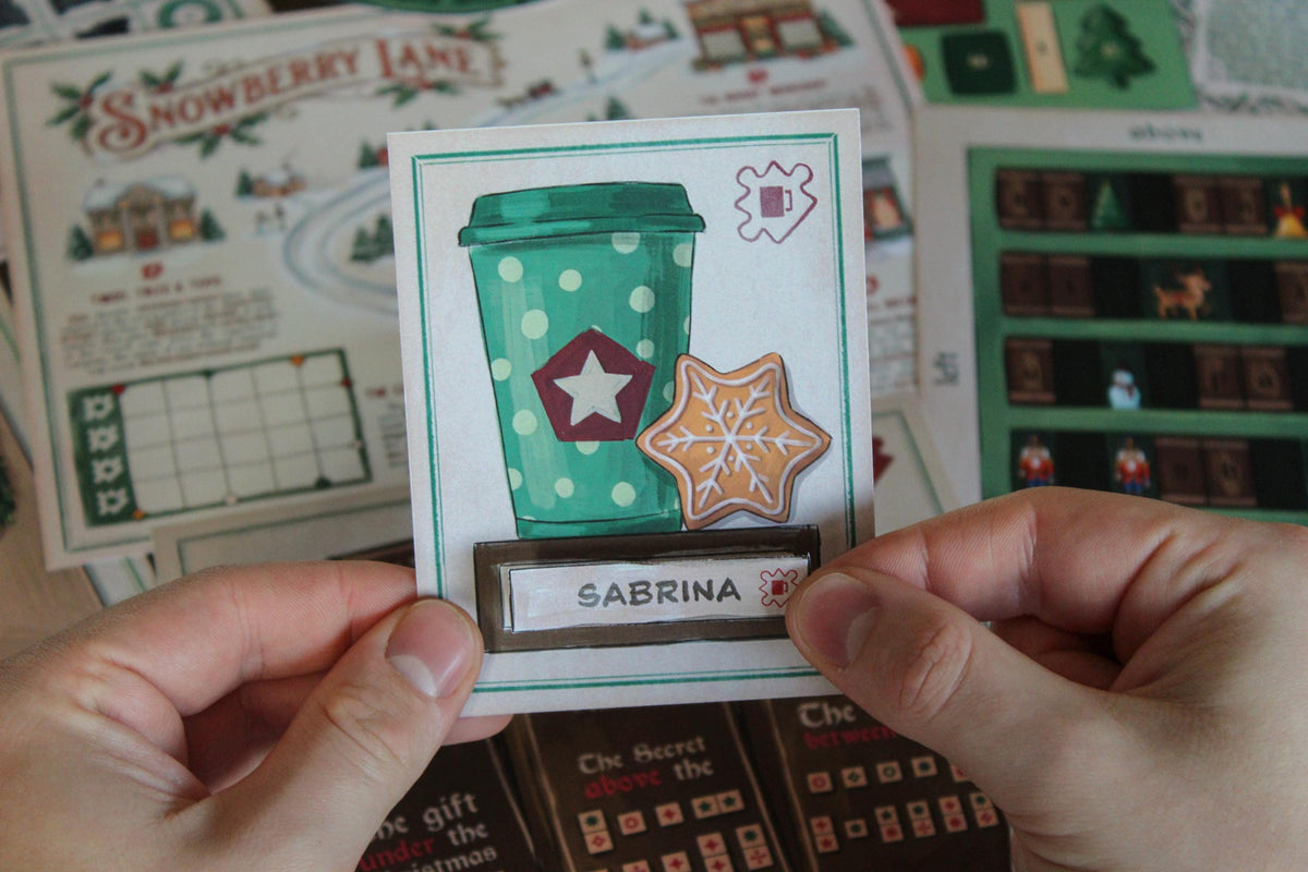 Close-up of hands holding a Cozy Christmas printable escape room puzzle card featuring a green polka-dot takeaway cup, a star-shaped gingerbread cookie with snowflake icing, and the name Sabrina. In the background are illustrated Snowberry Lane game materials, including the village map, answer grid, festive bookcase puzzle, Christmas books, and other printable components from the MysteryLocks Cozy Christmas Escape Room.