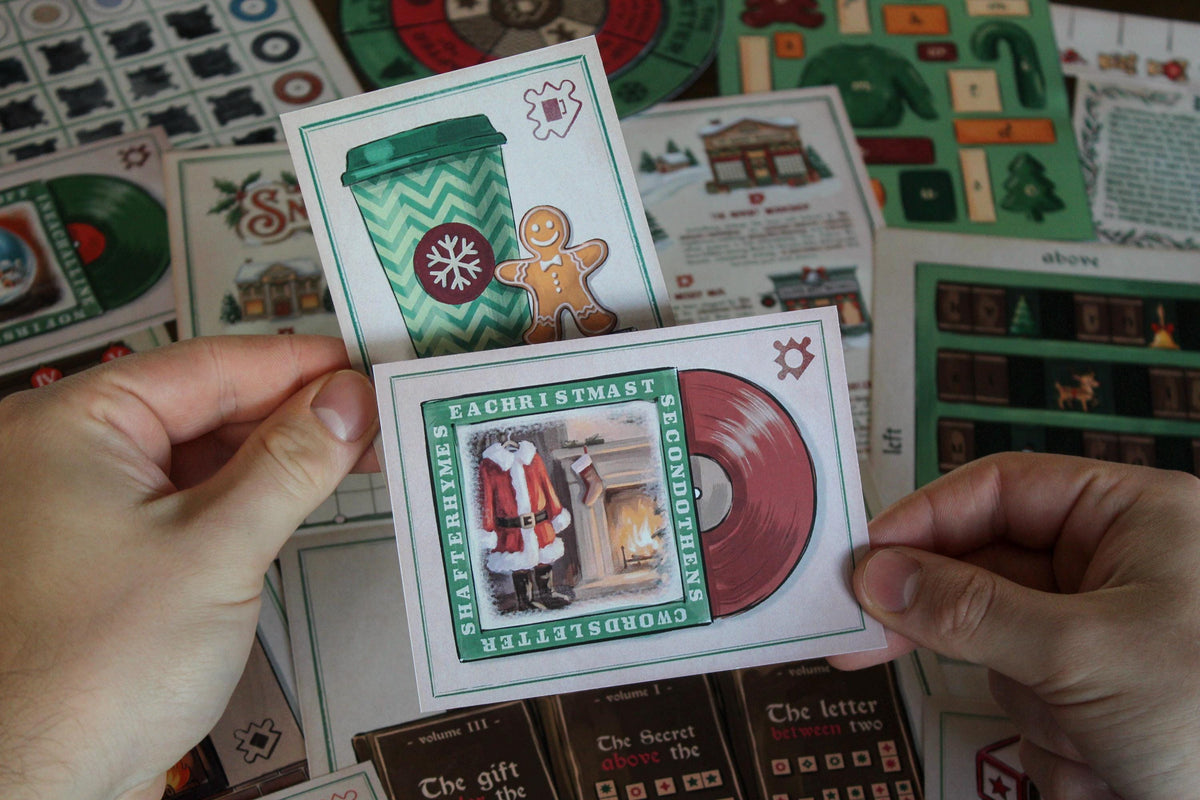 Close-up of hands holding two Cozy Christmas printable escape room puzzle pieces: a green takeaway cup with a gingerbread man and a red Christmas record featuring Santa beside a fireplace. In the background are illustrated Snowberry Lane materials including the village map, rotating decoder wheels, Advent Workshop pieces, bookcase puzzle, lyric sheets, and other festive components from the MysteryLocks Cozy Christmas Escape Room.